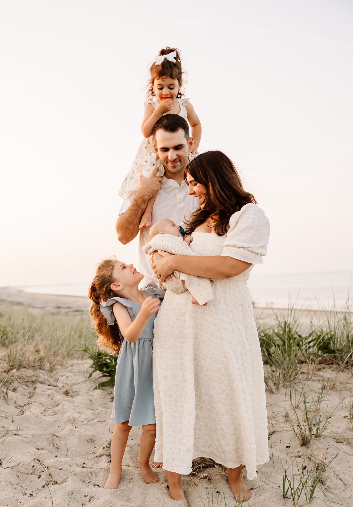 Family of five posing on sandy beach at sunset, parents holding baby while daughters play around them.