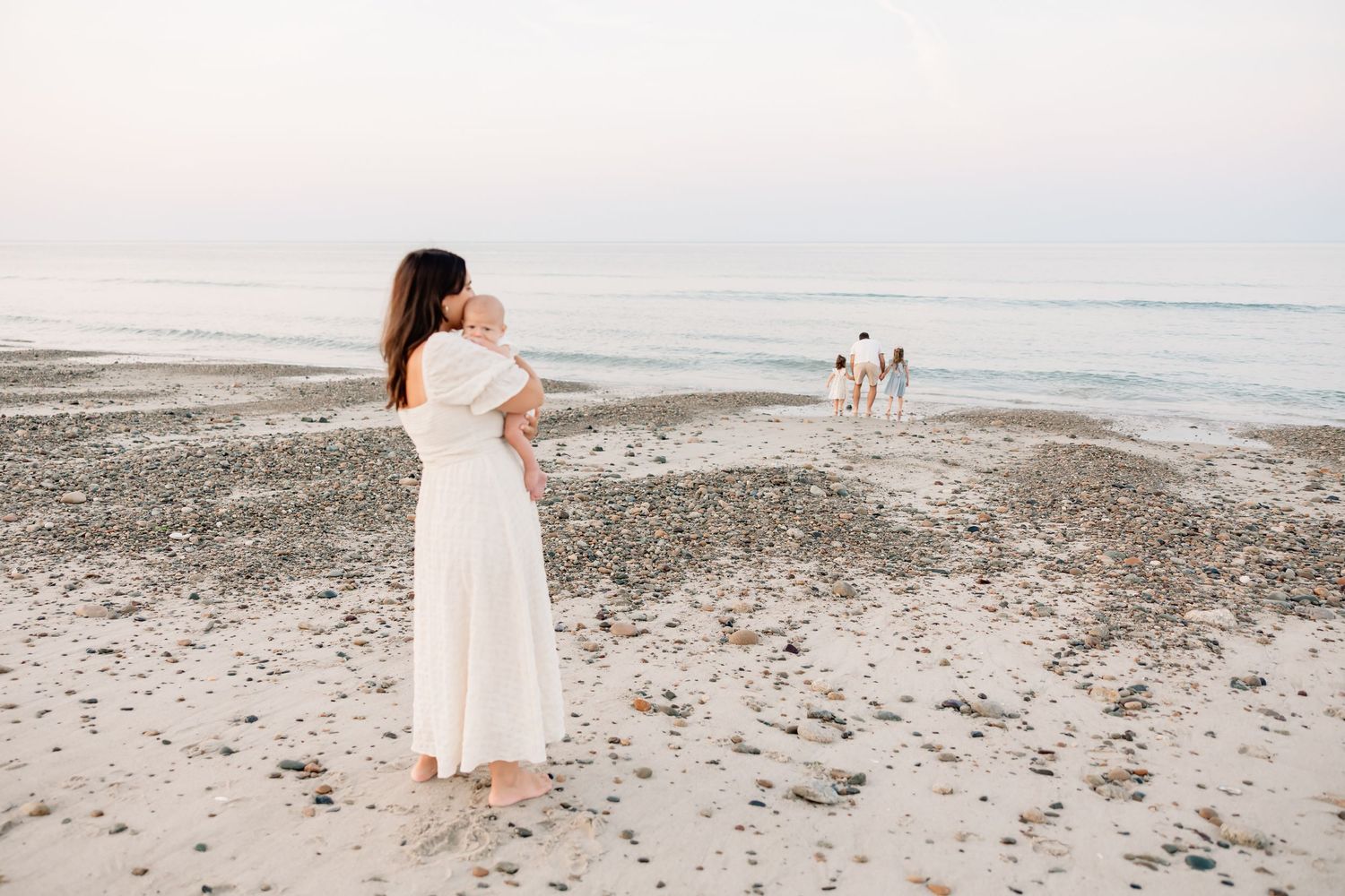 A mother in a white dress holds her baby on a serene beach at sunset with family in the background.