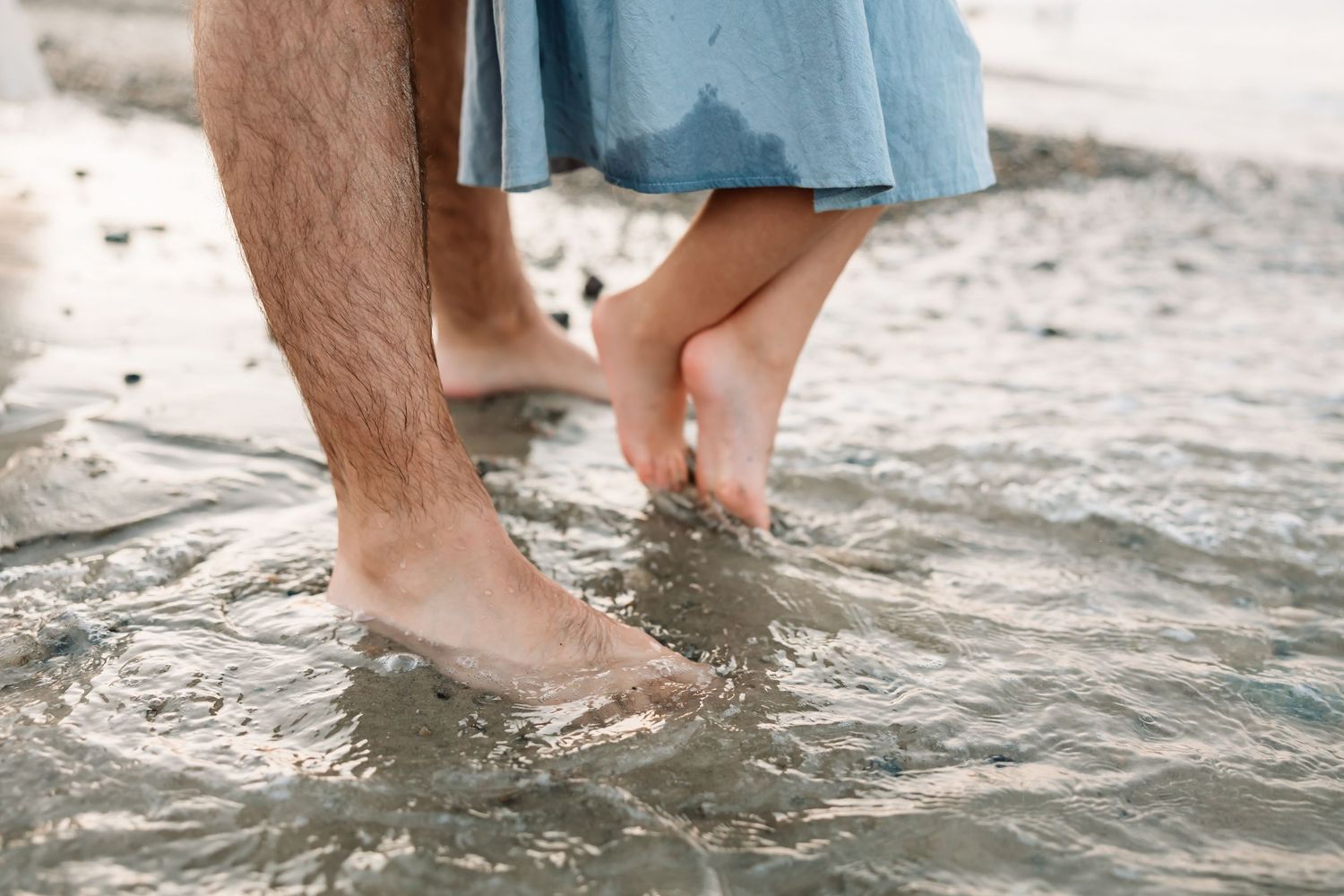 Couple standing barefoot in shallow ocean water, man in shorts and woman in blue dress on sandy beach.