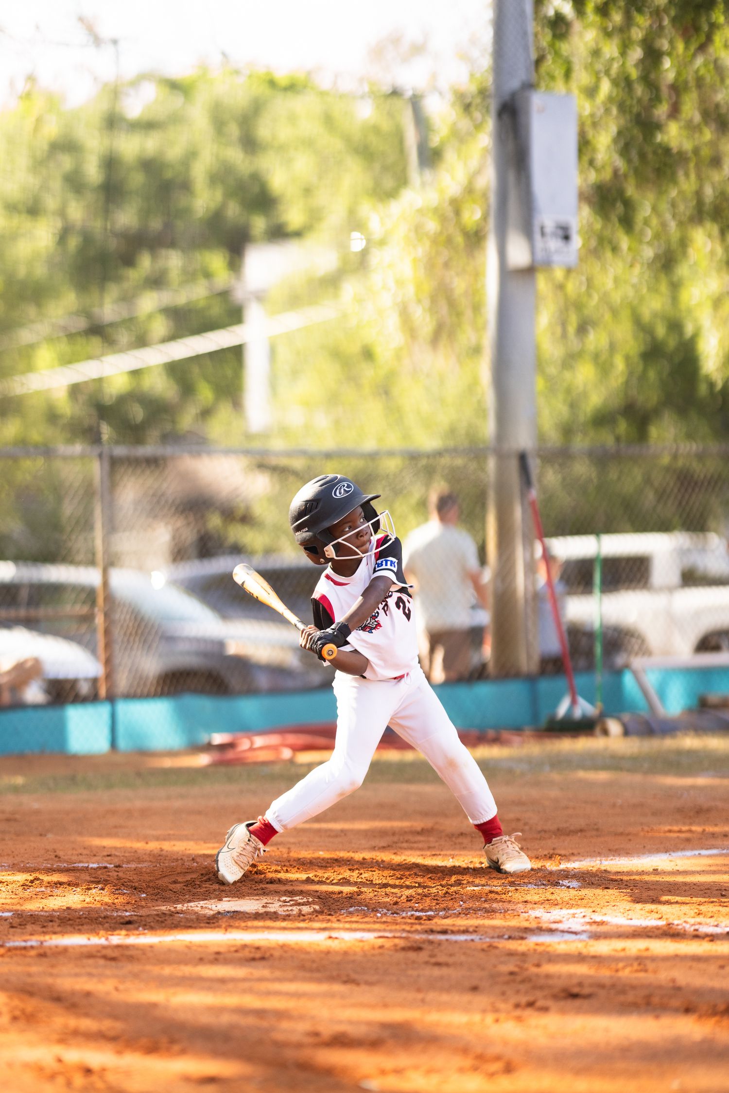 Young baseball player in white uniform swinging bat at home plate during a little league game on a dirt field.