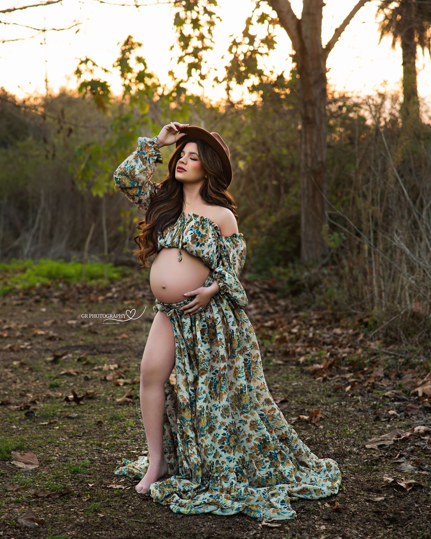 Pregnant woman in floral off-shoulder gown and brown hat poses outdoors at golden hour in a nature setting.