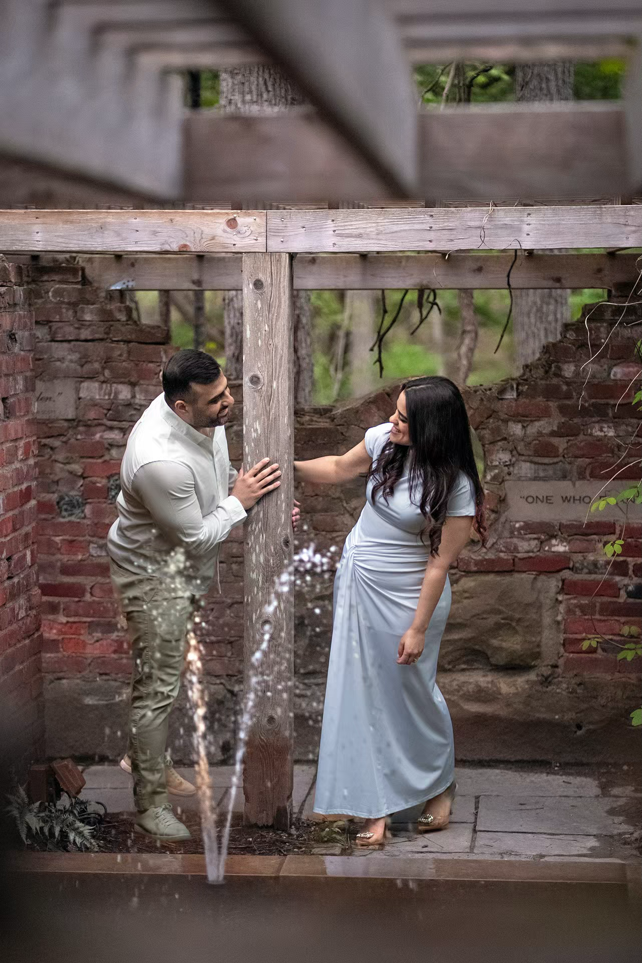 A couple shares a romantic engagement moment under a rustic wooden pergola with brick walls at inniswood Metro Gardens