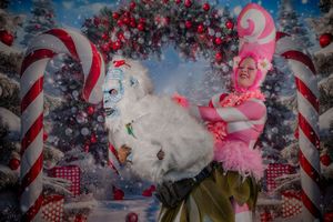 Two performers in colorful Christmas costumes pose in front of a festive holiday backdrop with candy canes and ornaments.