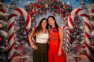 Two women smiling together in front of a festive Christmas backdrop with candy canes and decorated trees.