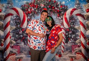 Couple posing in festive Christmas shirts in front of a holiday backdrop with candy canes and ornaments.