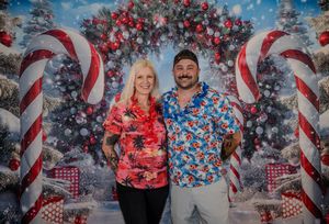 Couple in matching Christmas Hawaiian shirts posing before festive backdrop with candy canes and holiday decorations.