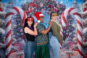 Three women pose in front of a festive Christmas backdrop with candy canes, wearing holiday accessories including a Santa hat.
