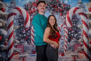 A couple poses together in front of a festive Christmas backdrop with candy canes and decorated trees.