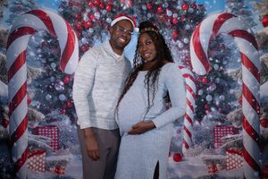 Smiling pregnant couple posing in front of festive Christmas backdrop with candy canes and decorated tree.