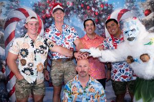 Five men in tropical Christmas shirts pose with a yeti mascot in front of a festive holiday backdrop.