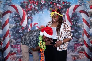 Two people pose in front of a festive Christmas backdrop with candy canes, one wearing a Santa hat and lei.