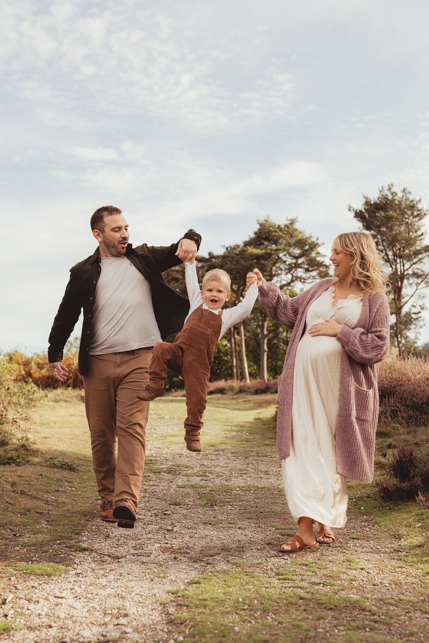 A happy family walks along a scenic heathland path, with dad carrying child on shoulders beside pregnant mom.