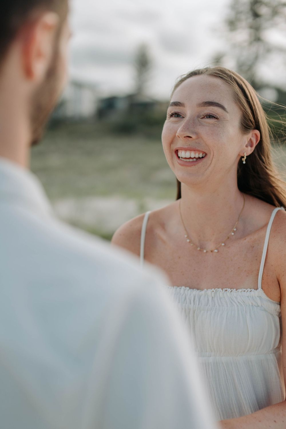 Smiling bride in white dress laughing joyfully during outdoor engagement or wedding ceremony.