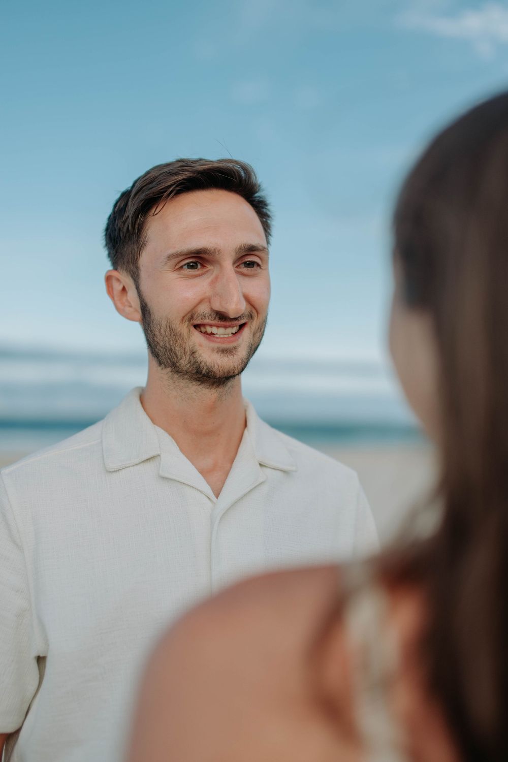 Smiling man with beard in white shirt at beach, looking at woman in foreground with blue sky background.