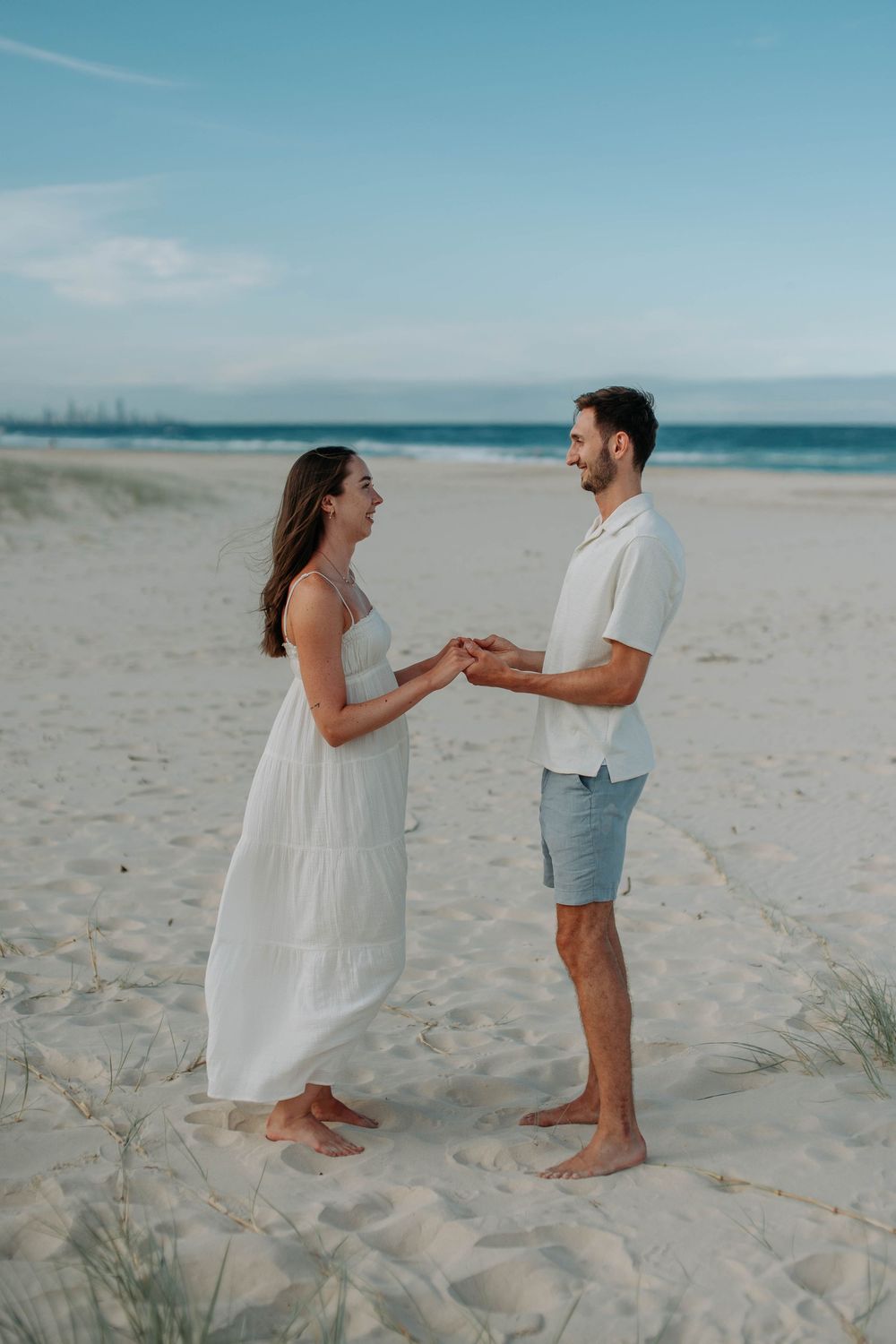 Couple holding hands on sandy beach, woman in white dress and man in white shirt and blue shorts.