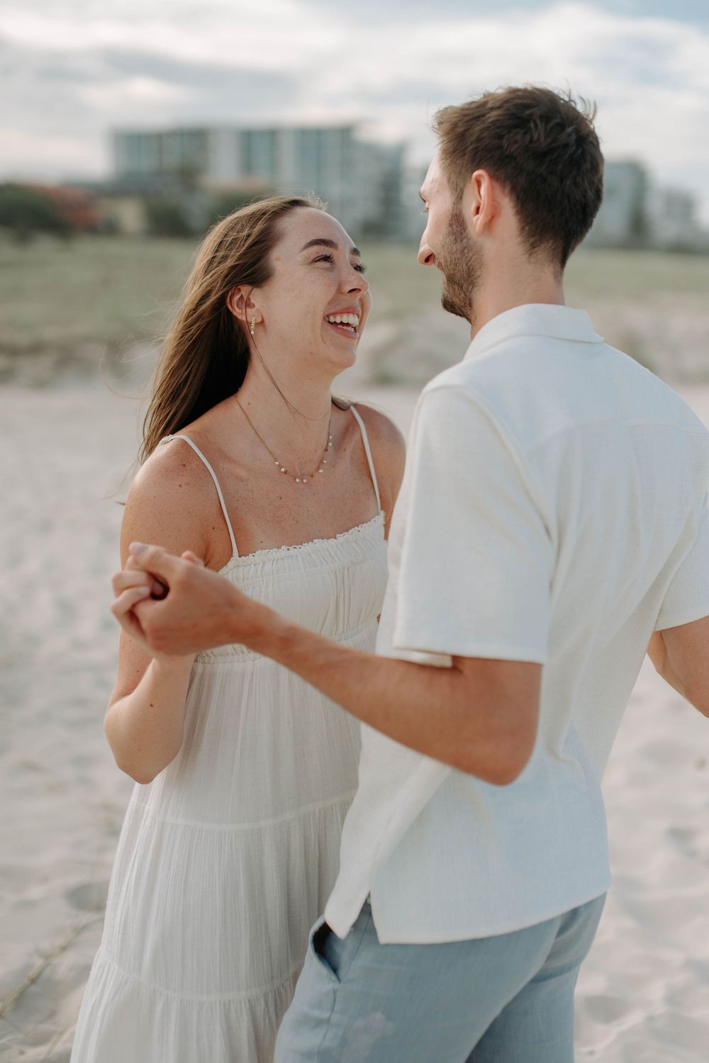 Happy couple dancing on sandy beach, woman in white dress laughing joyfully while holding hands with man in white shirt.