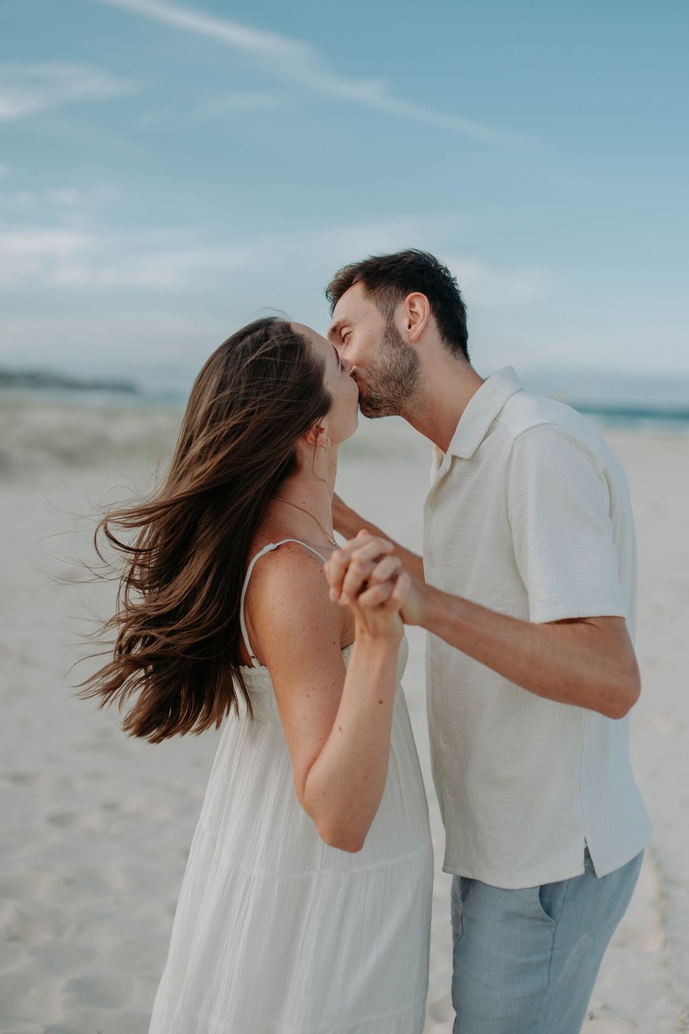 A couple shares a tender kiss while holding hands on a sandy beach, the woman's hair blowing in the wind.