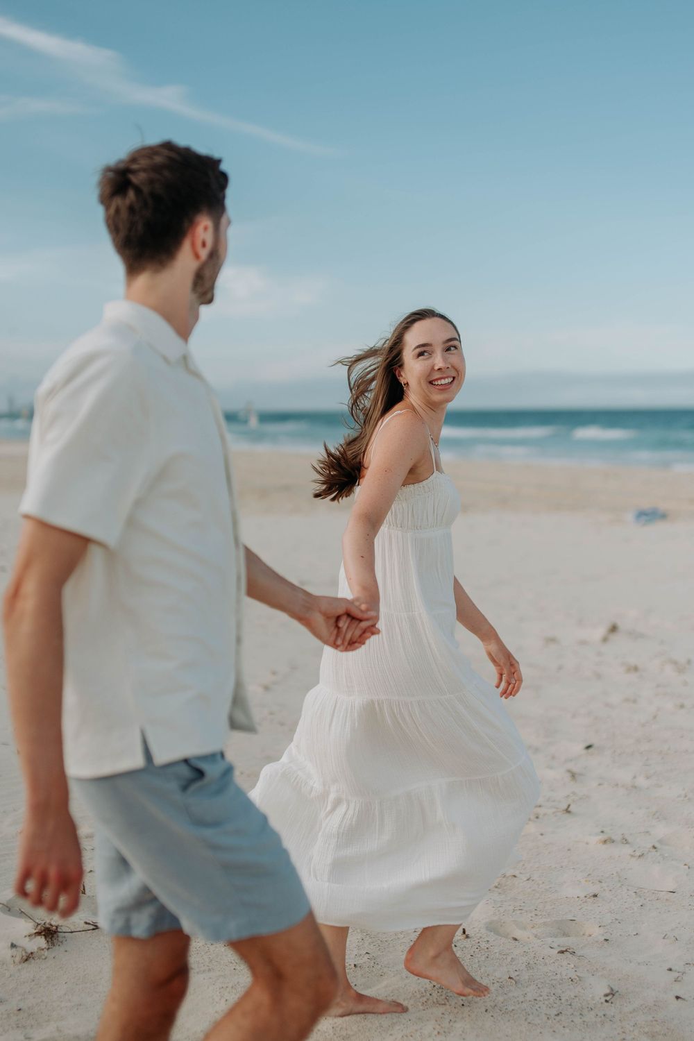 Happy woman in white dress walks hand in hand with man on sunny beach, hair blowing in breeze.
