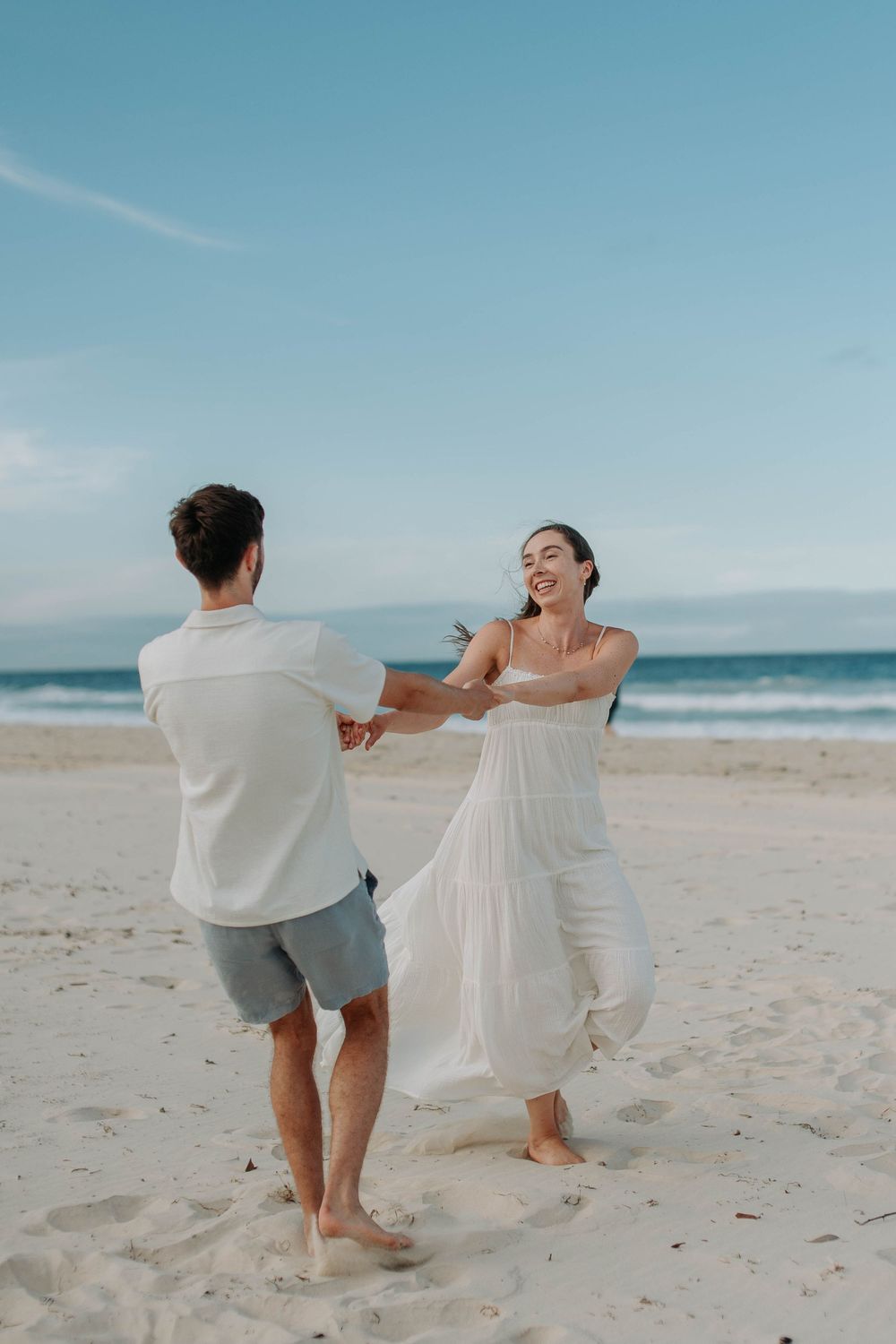 Joyful couple dancing together on sandy beach, woman in flowing white dress laughing with partner.