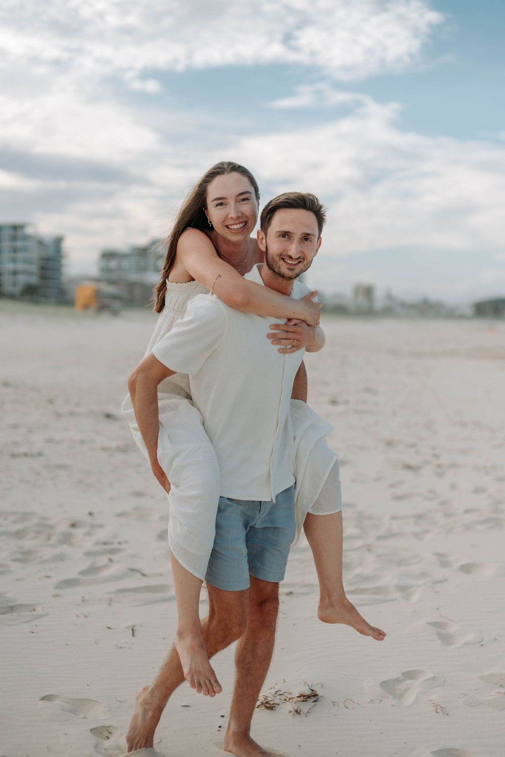 Smiling man gives woman a piggyback ride on sandy beach with buildings in background.
