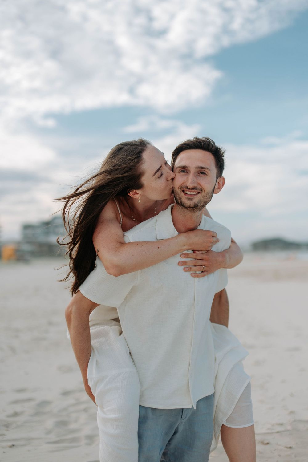 A smiling man gives his girlfriend a piggyback ride on the beach while she kisses his cheek.