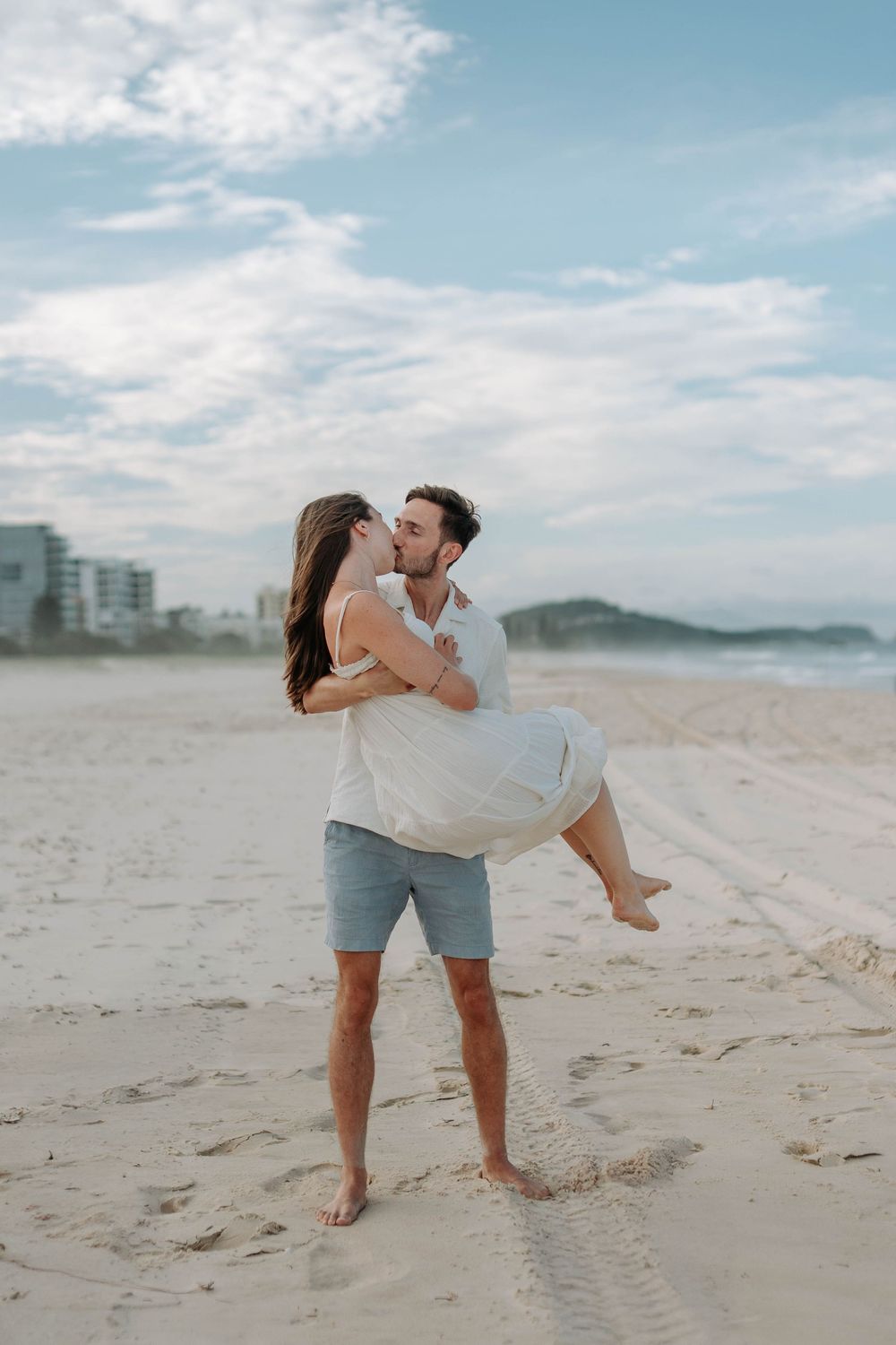 Man lifting woman in white dress while kissing on sandy beach with ocean backdrop.