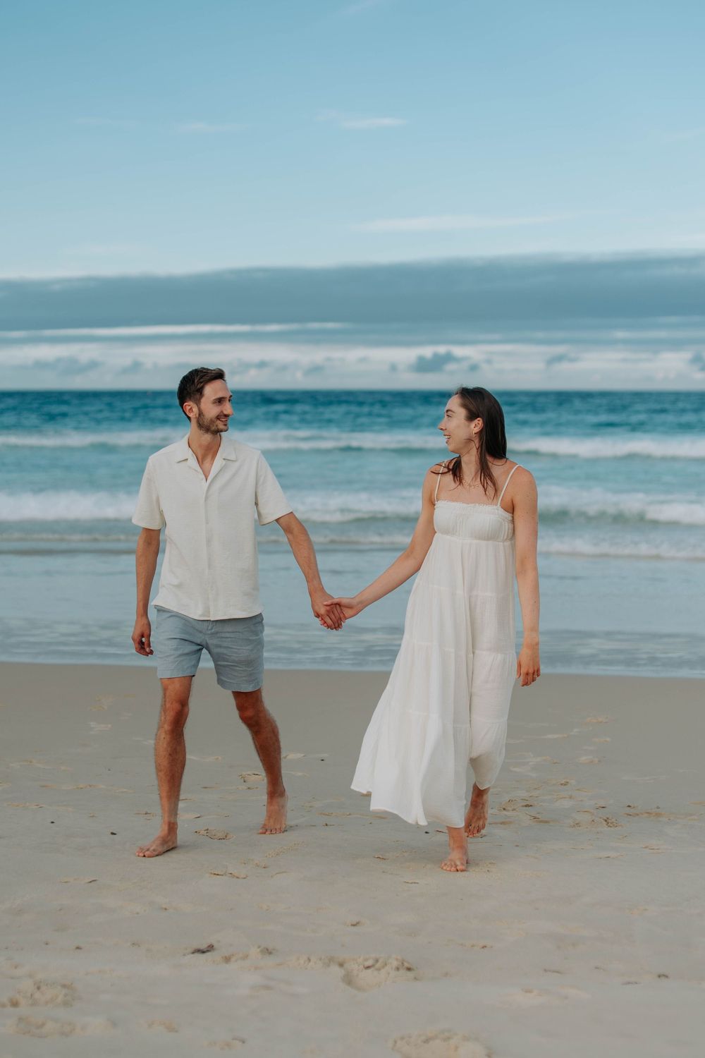 Couple holding hands and walking along sandy beach with ocean waves in background.