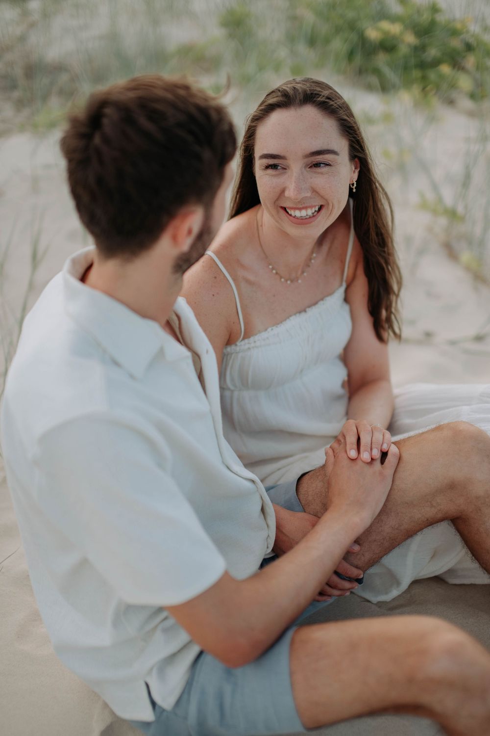 Smiling woman in white dress sits on sand dunes holding hands with man during beach photoshoot.