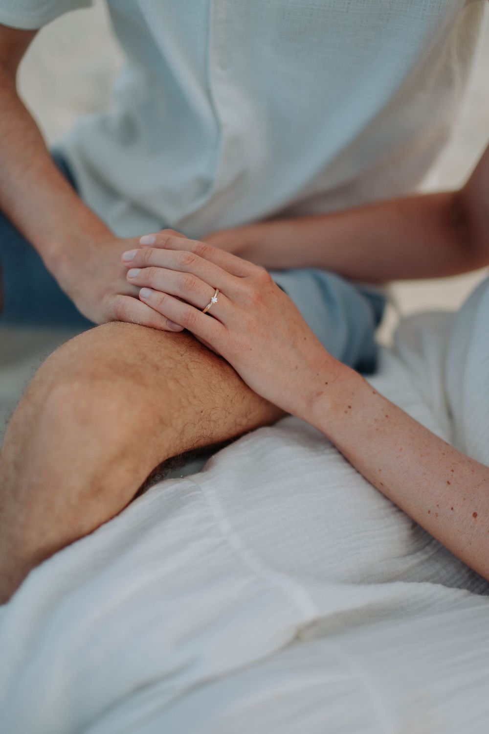 Close-up of woman's hand with gold ring resting on man's knee at beach.