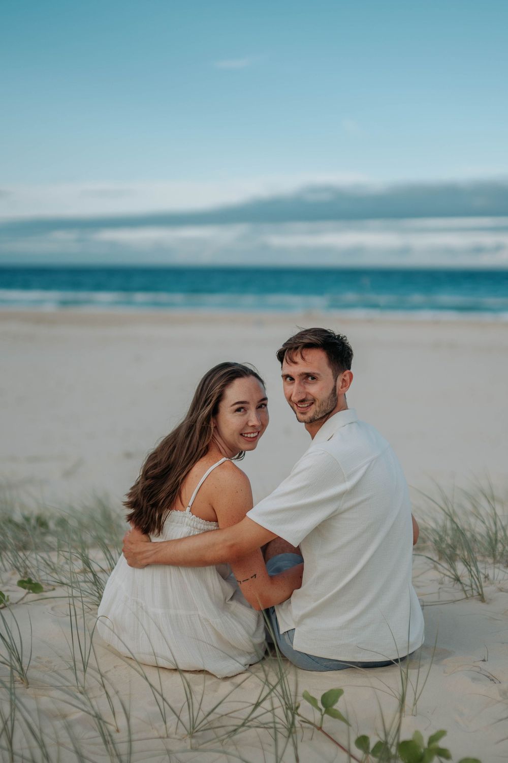 Couple sitting on sandy beach dunes, embracing and smiling, with turquoise ocean waves in the background.