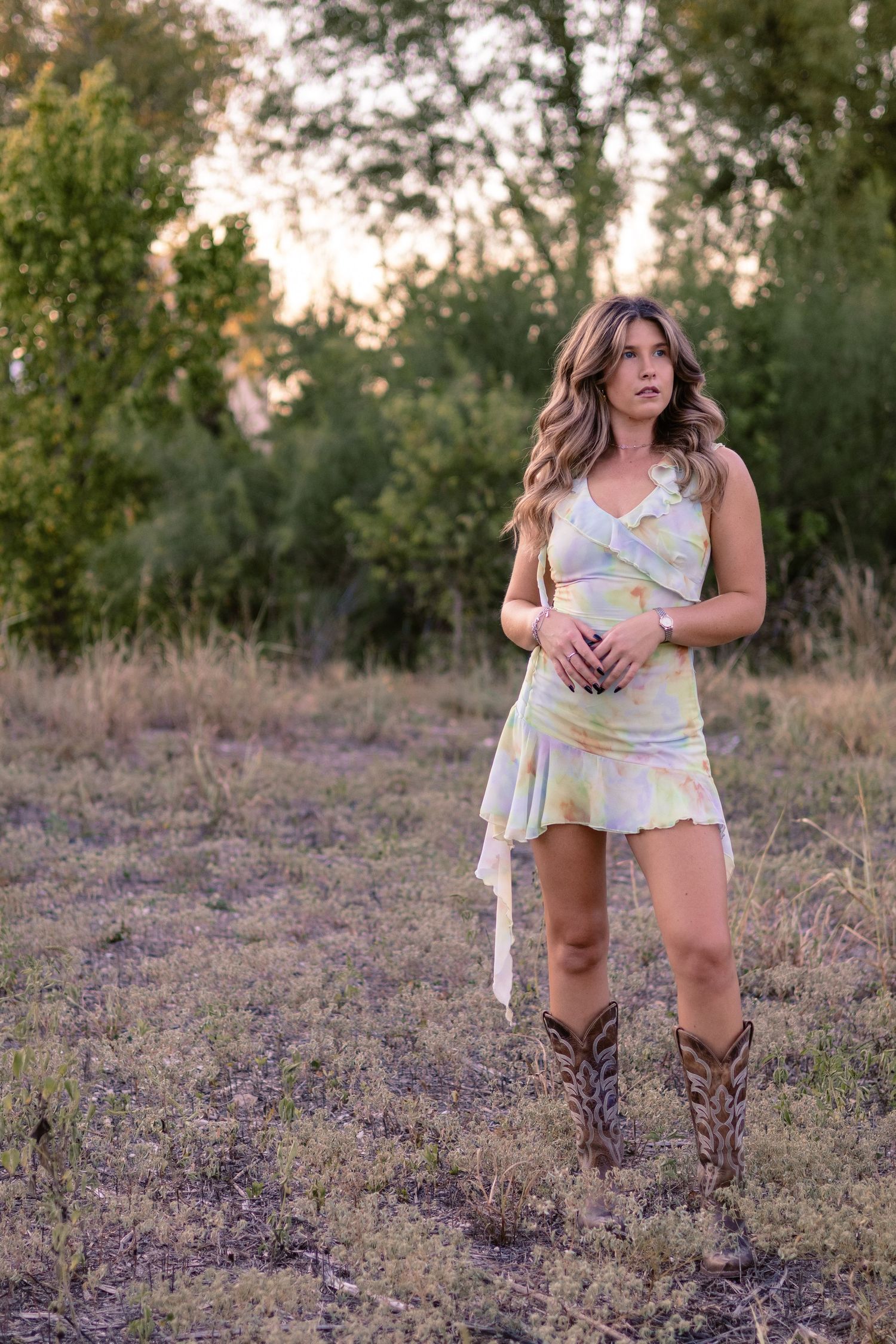 A natural light lifestyle portrait of a woman in a floral dress standing in an open Central Texas field during golden hour.
