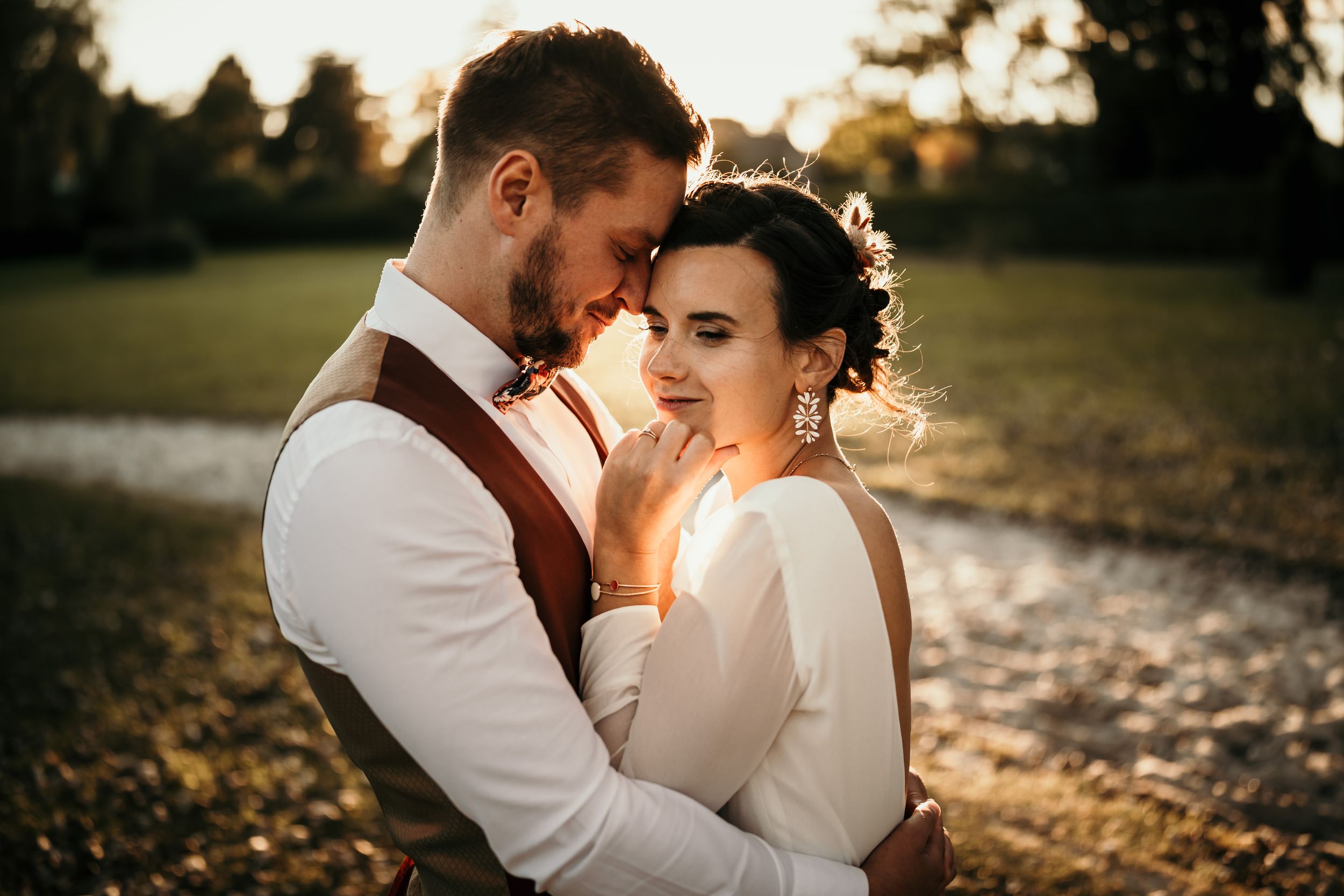 Romantic wedding couple embracing at golden hour sunset, bride in white gown with floral hair accessory, groom in vest and tie.