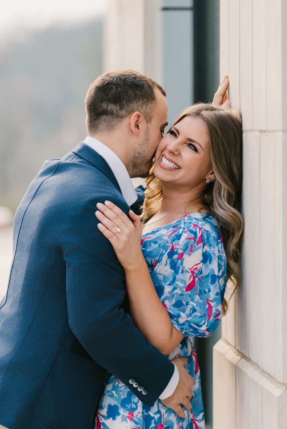 Smiling woman in floral dress being kissed by man in blue suit outdoors. Engagement session Bridge Park Dublin Ohio.