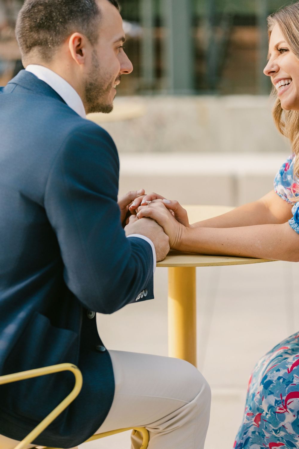 Engaged couple holding hands and smiling at a outdoor cafe table, man in navy suit, woman in floral dress.