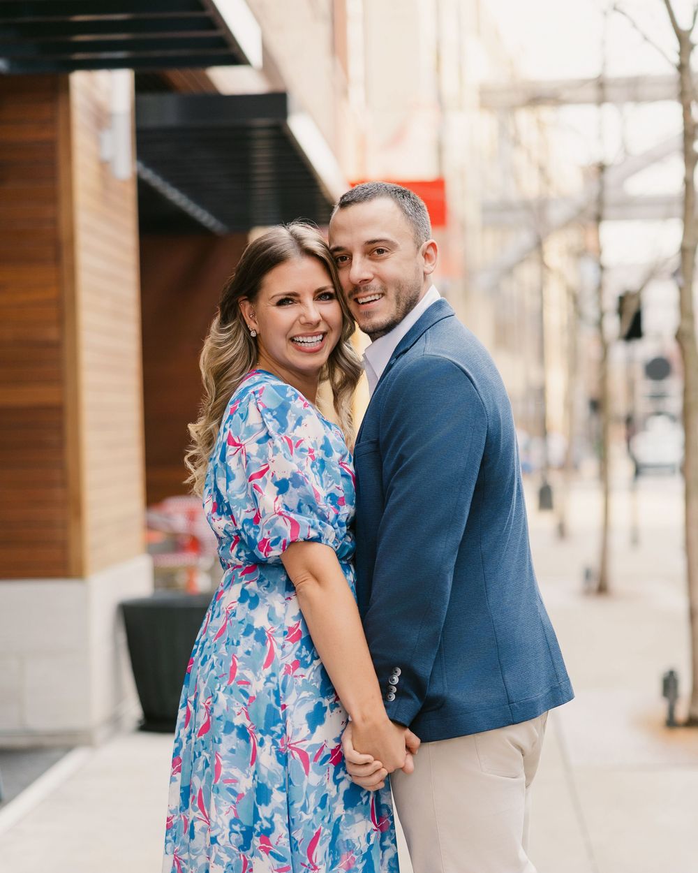A smiling couple poses on a city sidewalk, woman in floral blue dress and man in navy blazer with khaki pants.