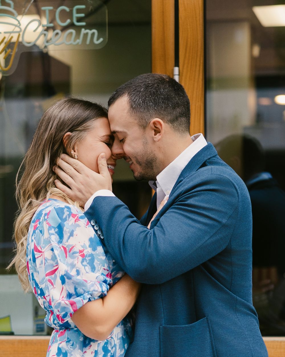 Couple sharing a romantic moment, foreheads touching, man in blue suit and woman in floral dress outside an ice cream shop.
