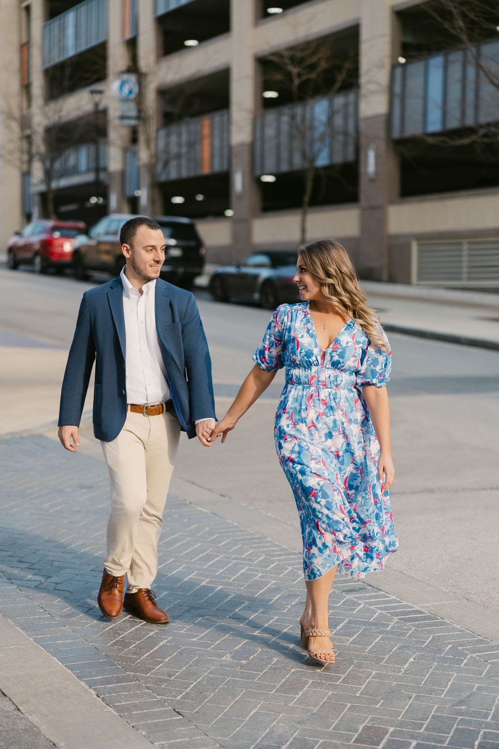 Couple holding hands and walking on a brick sidewalk, man in navy blazer and woman in blue floral dress.