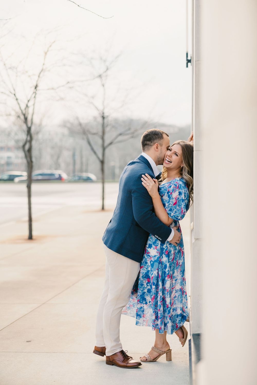 Couple embracing and laughing against a white column outdoors, man in navy blazer, woman in blue floral dress.