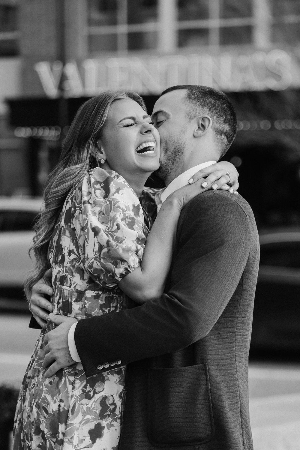 Black and white photo of a joyful couple embracing and laughing outside a Valentino store during an engagement session.