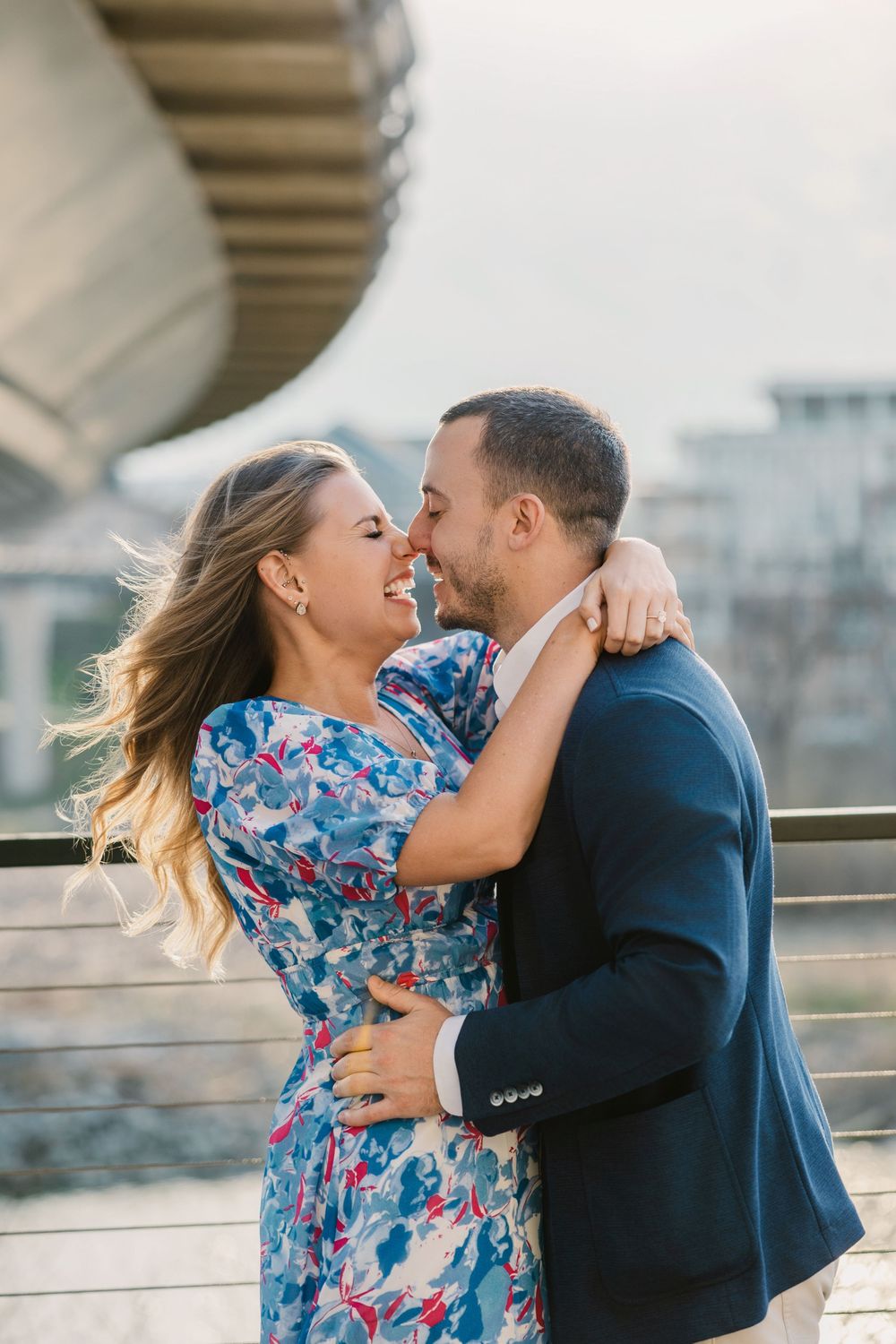 A couple embraces and smiles at each other outdoors, woman in floral dress, man in navy blazer, urban background.
