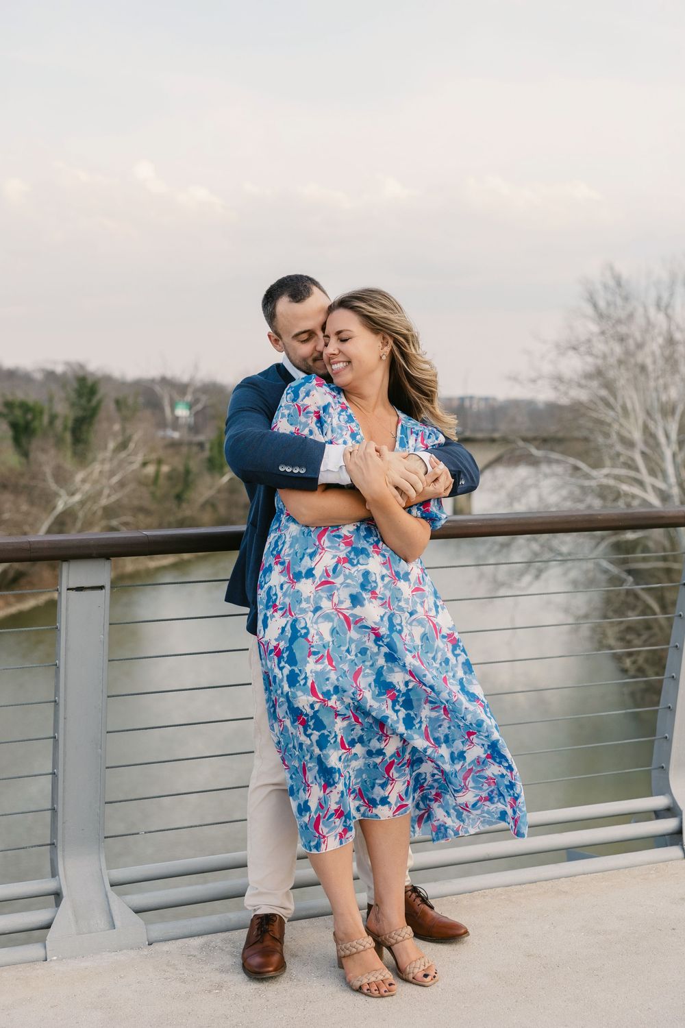 Couple sharing a romantic embrace on a bridge overlooking a river, woman in blue floral dress smiling joyfully.
