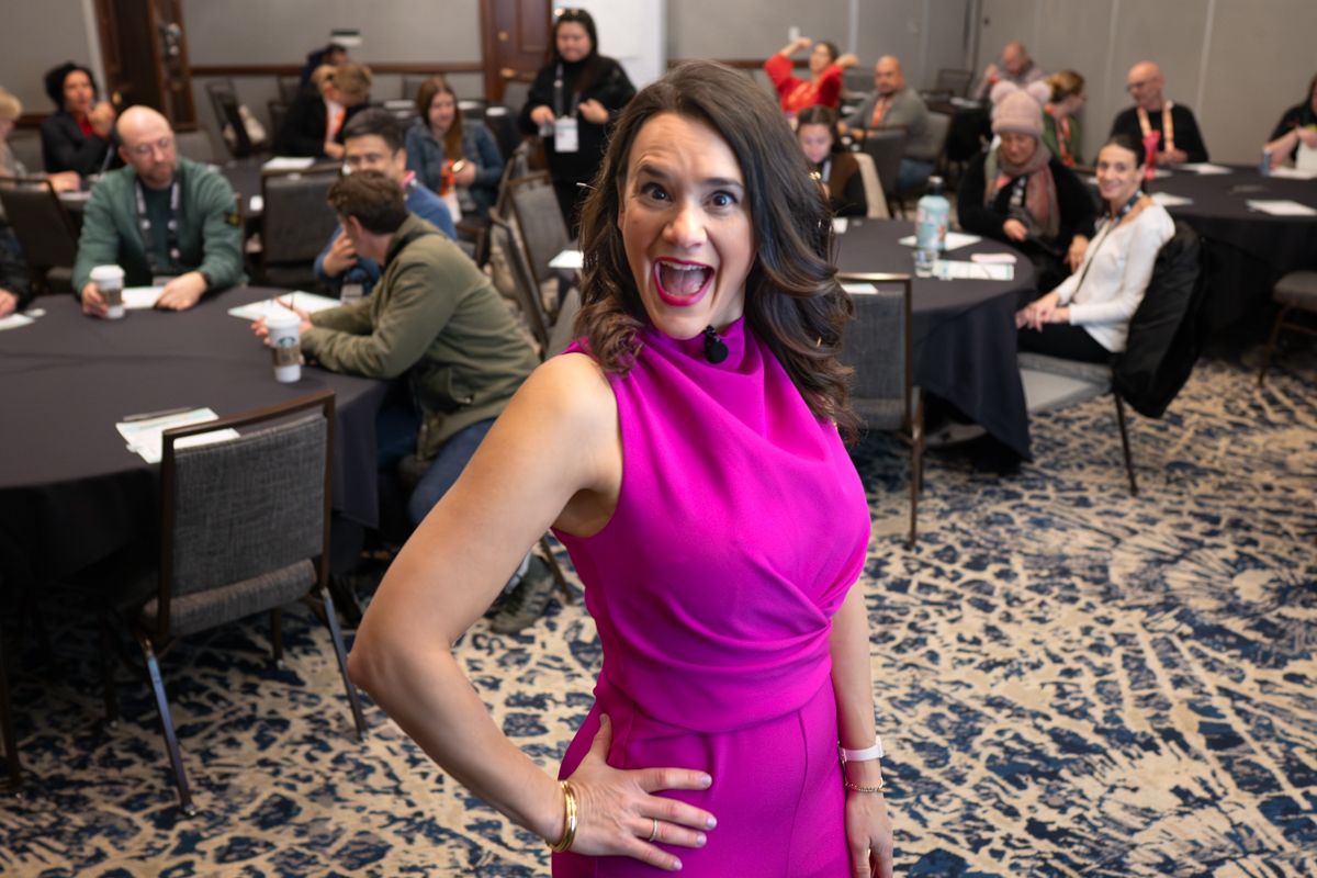 Woman in vibrant magenta dress poses confidently at a conference event with attendees seated at round tables behind her.