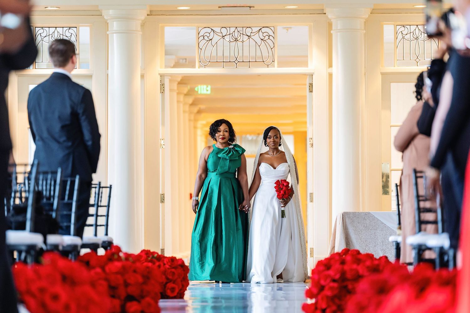 Bride walks down aisle with bridesmaid in teal gown, flanked by red roses, in elegant white wedding venue. Deerfield Golf Club Newark De