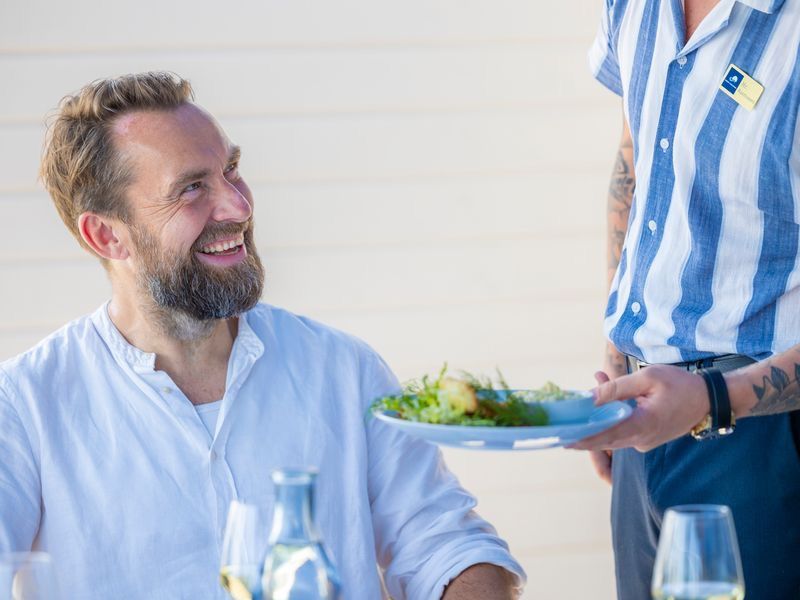 Smiling bearded man being served a fresh green salad by a waiter in a striped shirt at an outdoor restaurant.