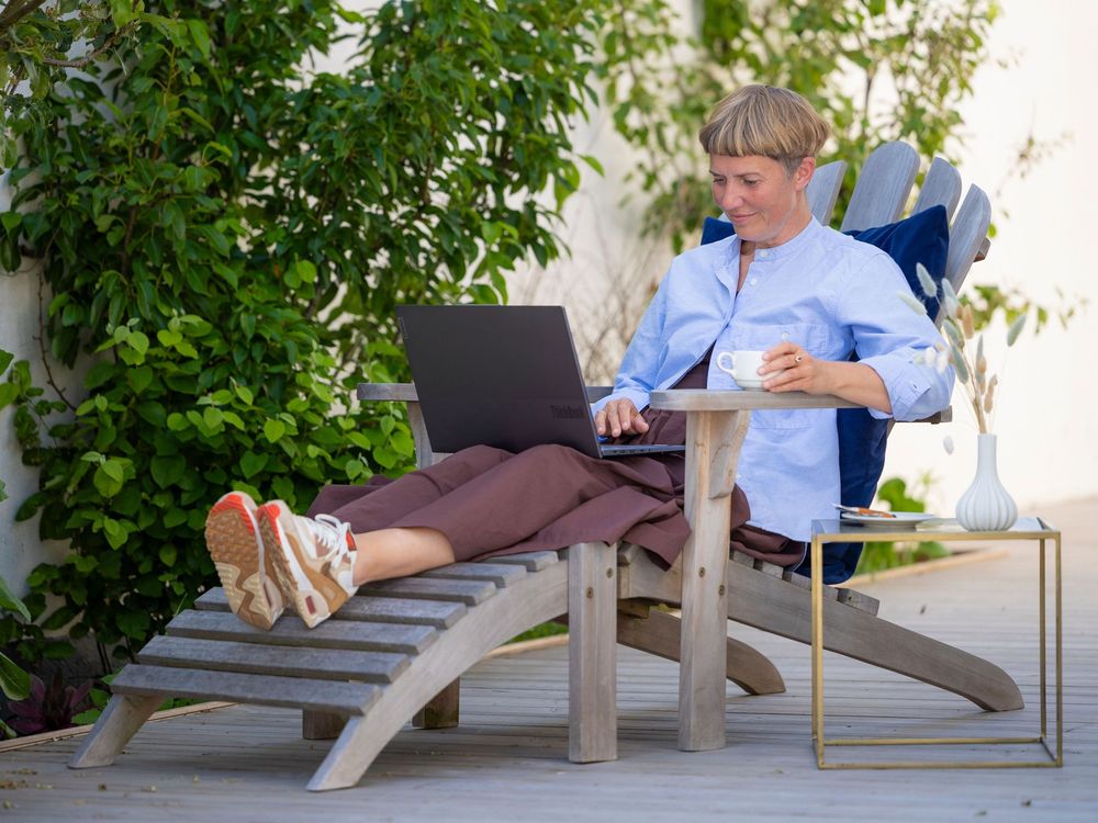 Person relaxing outdoors on wooden Adirondack chair with laptop and coffee cup, surrounded by lush green plants.