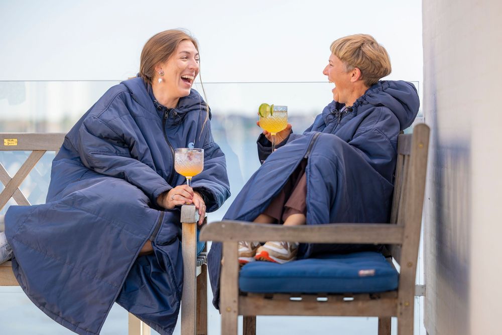 Two women laughing and holding drinks while wrapped in navy blue wearable blankets on an outdoor patio.