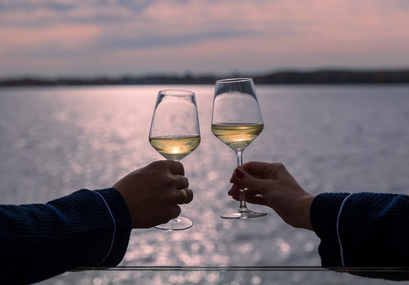 Two hands clinking glasses of white wine at sunset over a calm lake during a romantic evening.