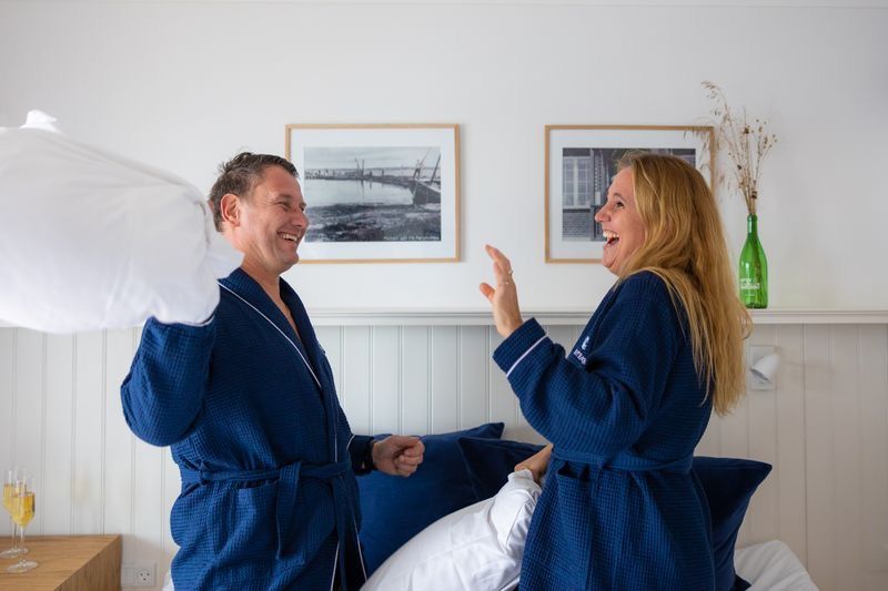 Couple laughing in matching navy blue robes having a pillow fight in a bright hotel bedroom.