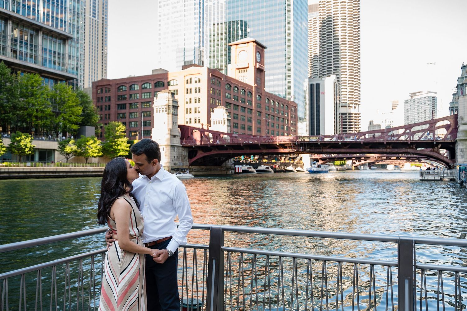 Couple embracing by Chicago River with city bridges and skyline in the background during golden hour.
