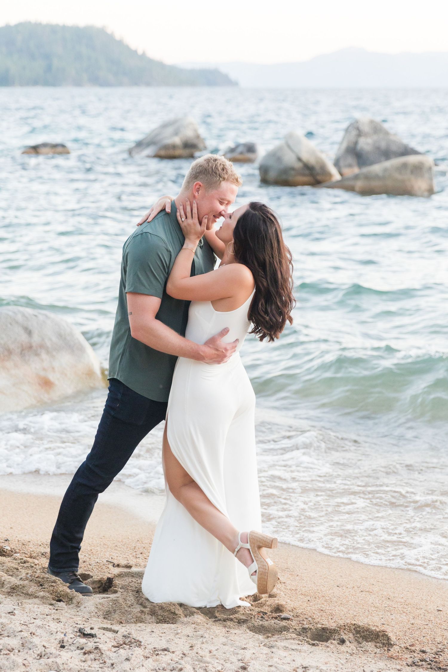 Couple sharing a romantic moment on a sandy Lake Tahoe beach surrounded by pine trees during golden hour.
